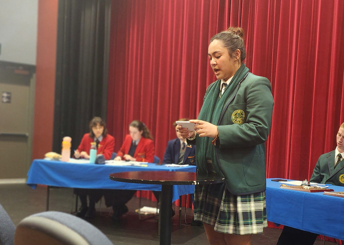 Mercedes College student confidently delivering a speech during an interschool debate, standing in front of a red curtain with adjudicators and competitors in the background.