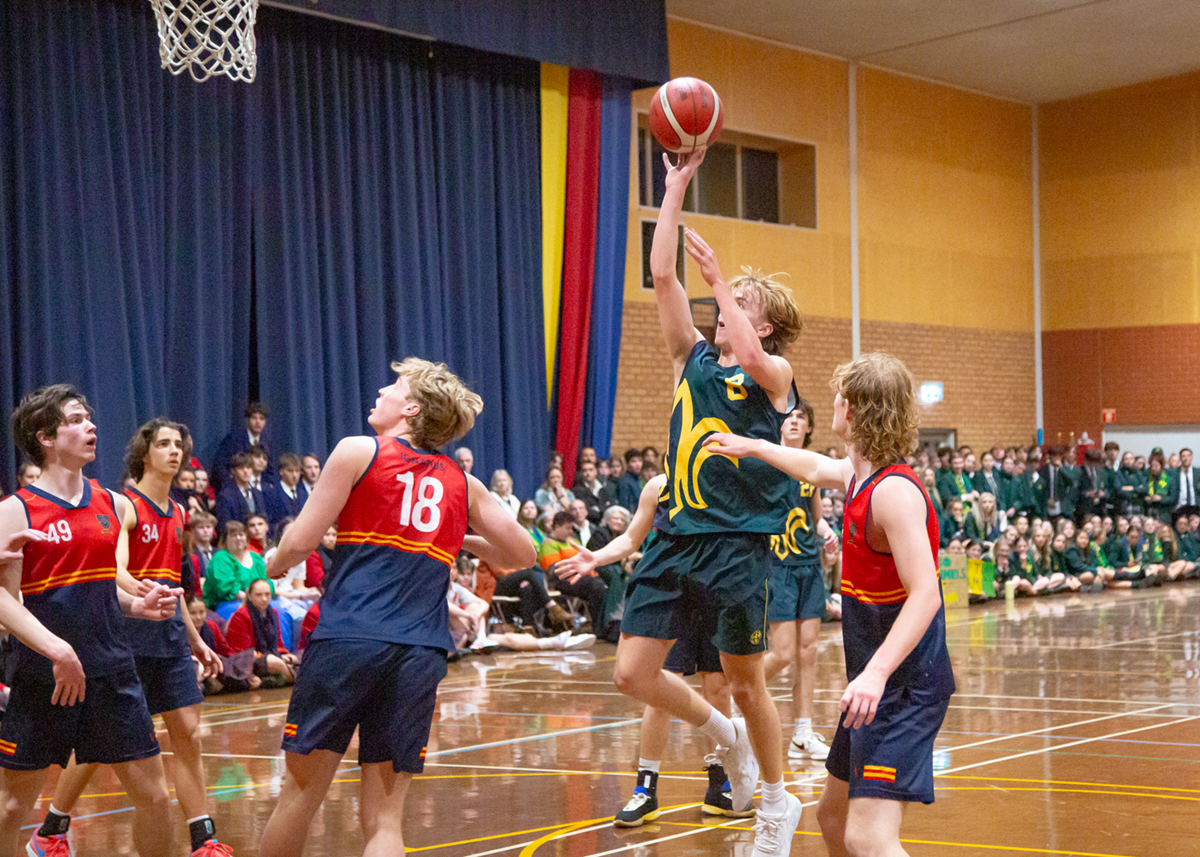 Mercedes College student jumps to shoot a basketball during an interschool match, with a packed crowd of students watching from the sidelines.