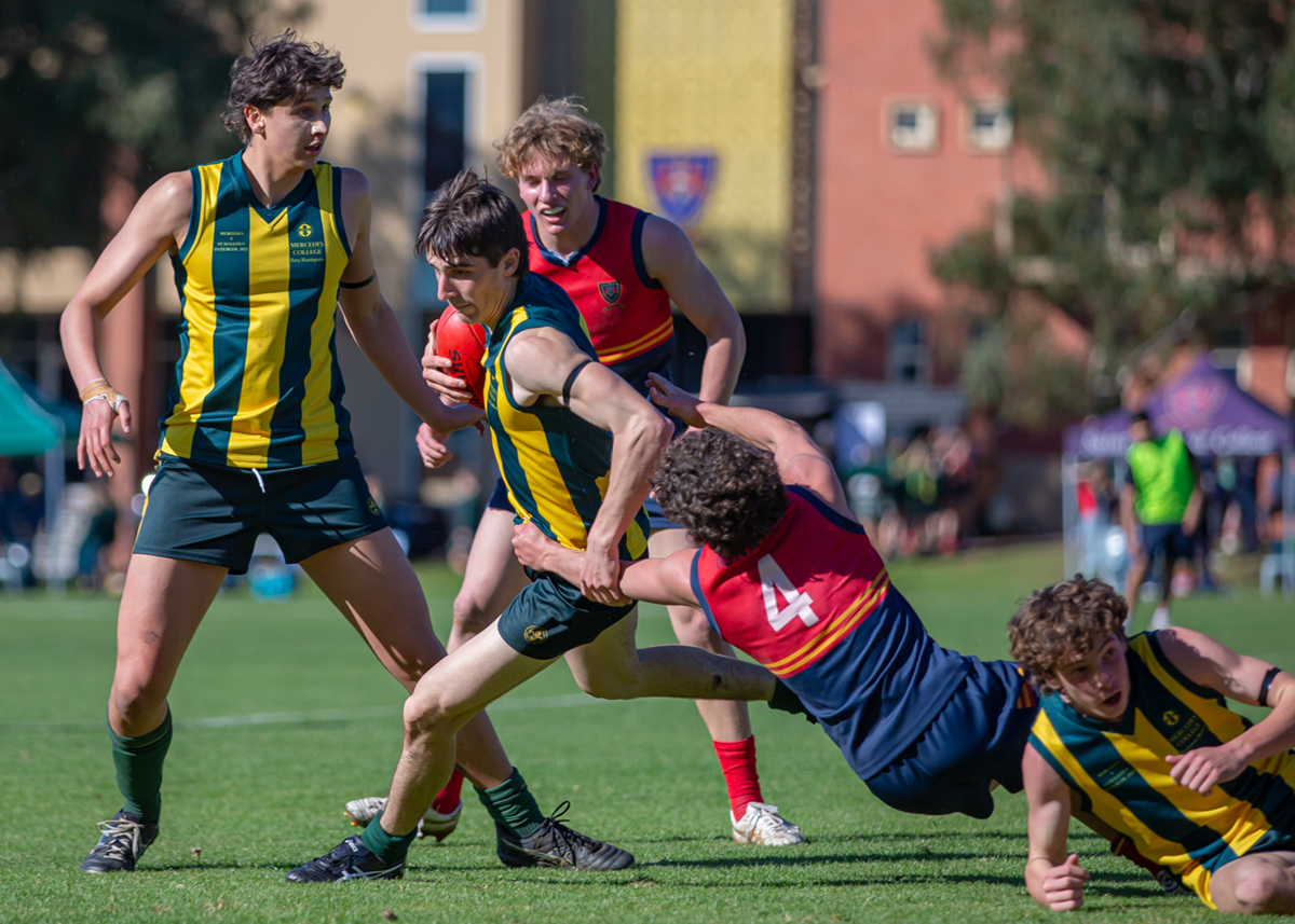 A fast-paced football match between Mercedes College and a rival school, featuring student athletes in mid-play on the school oval.