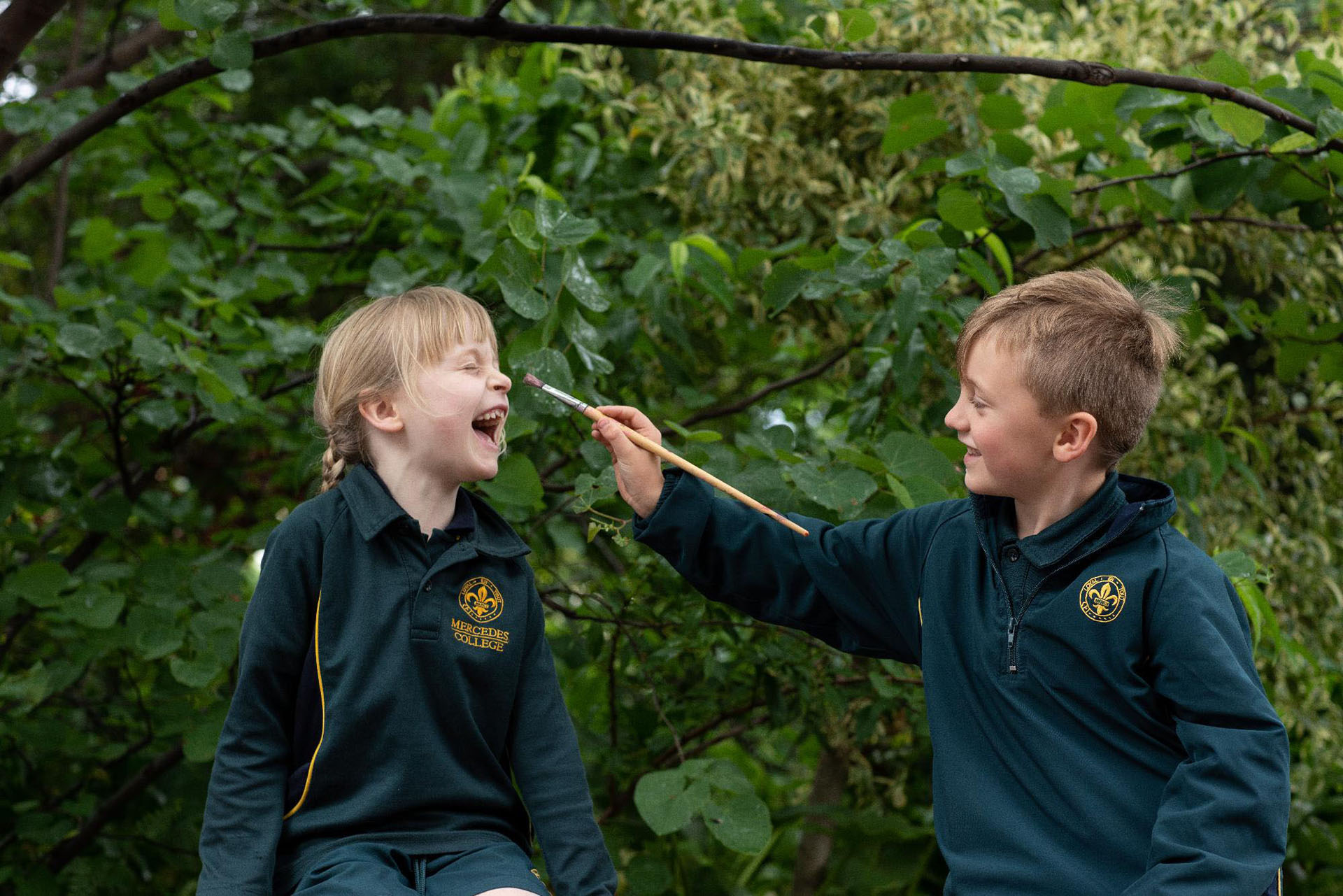 Young Mercedes College students dressed in green school uniforms, having fun as one paints the other's face during an outdoor activity.