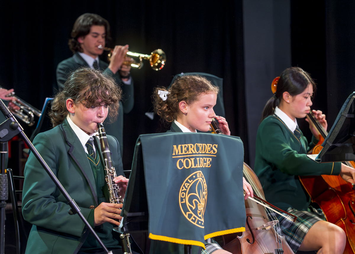 Mercedes College students in formal uniform performing in a school band, playing clarinet, cello, and trumpet on stage.
