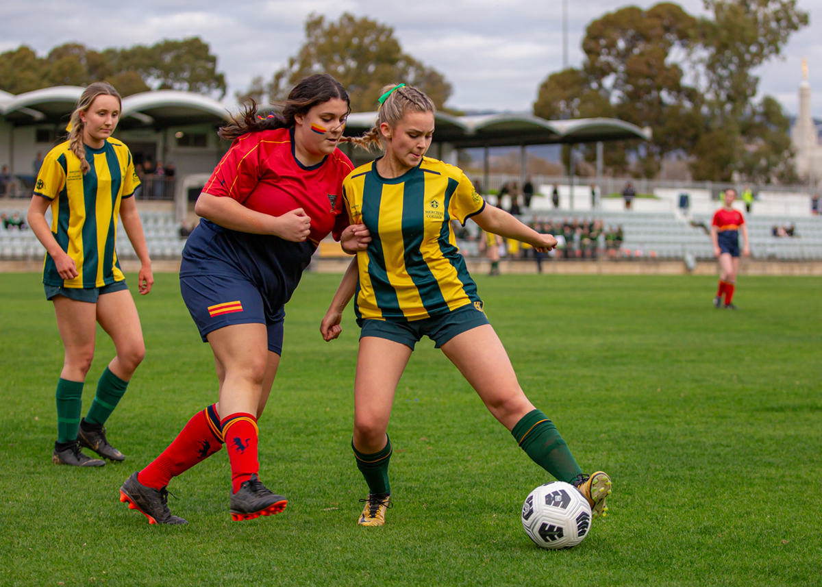 A Mercedes College student in green and gold uniform controls the soccer ball during a match, with teammates and opponents nearby on a grassy field.