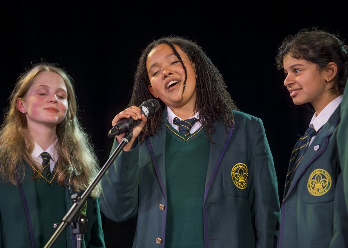 Student vocalist sings passionately into a microphone on stage at Mercedes College, accompanied by two fellow students in formal school uniforms.